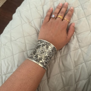 May include: A silver cuff bracelet with an intricate floral design is worn on a person's wrist. Several rings adorn the fingers, including a ring with a large, oval-shaped stone and two gold bands.