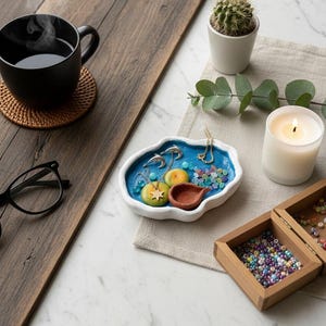 Op de afbeelding: Een serene stilleven met een zwarte mok met stoom, een kleine cactus in een witte pot en een brandende kaars. Een blauw-wit sieradenschaaltje bevat kettingen en decoratieve elementen. Houten dozen met kralen zijn ook aanwezig.