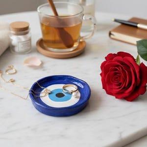 May include: A blue and white ceramic trinket dish with an eye design holds jewellery. A red rose, a cup of tea, and a small notebook with a pen are also on the marble surface. A small jar and a folded towel are also present.