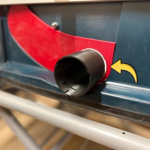 May include: Close-up of a table saw dust port. The saw features a dark blue body with a red safety guard. A black cylindrical dust port is visible, along with a yellow arrow indicating the direction of dust flow. The saw sits on a gray metal frame.