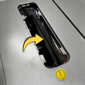 May include: A close-up of a silver table saw with a black blade guard and a yellow arrow sticker. A yellow circular sticker with text is in the lower right corner. The saw has a rectangular opening for the blade.