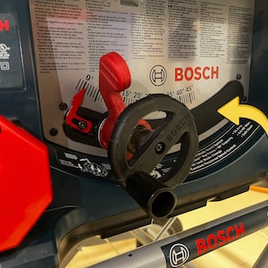 May include: A close-up of a Bosch 4100XC table saw. The saw features a red lever, a black handle, and a digital display. The saw has a blade guard system and a constant response speed control.