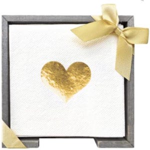 Op de afbeelding: Een vierkante stapel witte papieren servetten met een metallic gouden hartontwerp in het midden. De servetten zitten in een donkergrijze houten lijst en zijn versierd met een glanzend gouden lint dat in een strik in de rechterbovenhoek is gebonden.