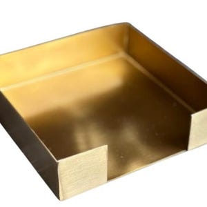 May include: A square, open-top metal tray with a brushed gold finish. The container features four vertical walls of equal height, with a U-shaped cutout on one side, creating a low-profile, box-like structure with a smooth, metallic surface.