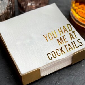 May include: A stack of square white paper napkins held in a gold-colored metal holder. The top napkin features the text YOU HAD ME AT COCKTAILS printed in a metallic gold foil font with a textured, slightly distressed appearance.