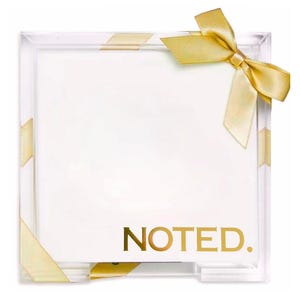 May include: A square clear acrylic holder containing a stack of white paper sheets. The word NOTED. is printed in gold lettering at the bottom right. A gold satin ribbon is tied in a bow at the top right corner of the acrylic frame.