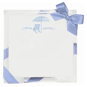 May include: A square stack of white paper sheets featuring a blue line drawing of a beach umbrella and a lounge chair. The stack is wrapped with a light blue satin ribbon tied in a bow at the top right corner and secured at the bottom edges.