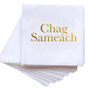 May include: A stack of square white paper napkins featuring the text "Chag Sameach" printed in a metallic gold font. The napkins have a subtle diagonal embossed line pattern across the surface, creating a textured appearance on the white paper.