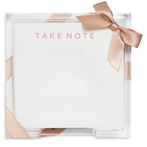 May include: A square clear acrylic holder containing a stack of white paper sheets with the text TAKE NOTE printed in light pink. The holder is wrapped with a light pink satin ribbon tied into a bow at the top right corner of the frame.