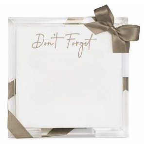 May include: A square clear acrylic memo holder containing a stack of white paper sheets. The text Don't Forget is printed in a light brown script font on the top sheet. A taupe-colored satin ribbon is tied around the corner of the acrylic frame.