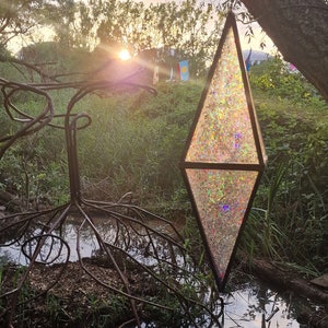 Pode incluir: Uma grande escultura suspensa em forma de diamante, iridescente, com uma bola de discoteca pendurada em um galho de árvore sobre um pequeno lago. A escultura é feita de vidro e reflete a luz em um arco-íris de cores.