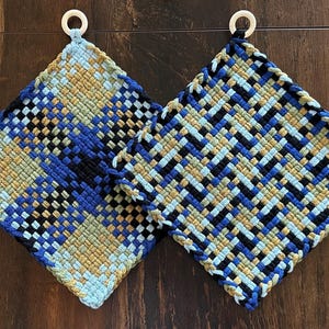 May include: Two woven potholders with a geometric pattern in shades of blue, black, and tan. Each potholder has a white ring for hanging. The potholders are square and appear to be handmade.