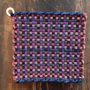 May include: A square, woven hot pad in a patchwork design. The pad features a mix of colors including blue, purple, green, pink, and brown. A white loop is attached to one corner for hanging. The edges are finished with a contrasting stitch.