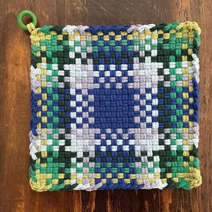 May include: Square, woven hot pad in shades of blue, green, yellow, and gray. The center features a darker blue square pattern. A green loop is attached to one corner for hanging. The hot pad is likely handmade.