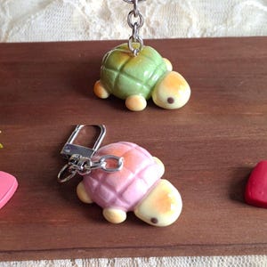 May include: Two turtle keychains featuring sculpted shells with a grid pattern. One turtle has a green shell with a pale yellow head, while the other has a pink shell with a pale yellow head. Both include a silver metal chain and a ring attachment.