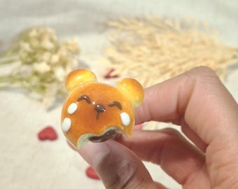 May include: A miniature bear-shaped pastry with a bite taken out, featuring a brown filling and white accents. The pastry has a glossy finish and is held in a hand. The overall theme is food and baked goods.