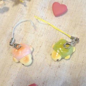 Op de afbeelding: Twee schildpadvormige bedels, één roze en één groen, met gele buikjes en bevestigde zilveren sluitingen. De bedels liggen op een lichtgekleurde stof, met roze hartvormige decoraties verspreid. Een geel koord is bevestigd aan de groene schildpadbedel.