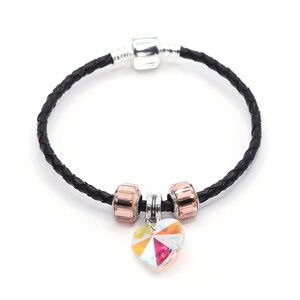 Könnte beinhalten: Ein schwarzes Lederarmband mit einem silbernen Anhänger und zwei roségoldenen Perlen. Ein Kristallherz-Anhänger hängt an den Perlen.