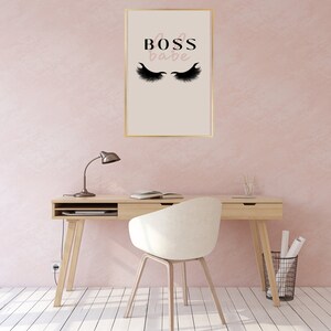 Op de afbeelding: Een ingelijst print met de tekst "Boss babe" in roze letters en een paar zwarte wimpers op een beige achtergrond. De print hangt aan een lichtroze muur boven een houten bureau met een witte stoel.