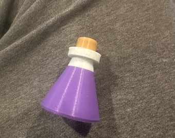May include: A small purple glass bottle with a white rim and a cork stopper. The bottle is sitting on a gray fabric surface.