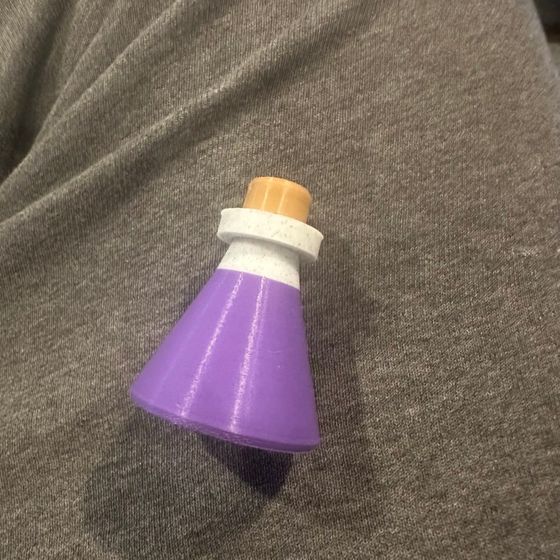May include: A small purple glass bottle with a white rim and a cork stopper. The bottle is sitting on a gray fabric surface.