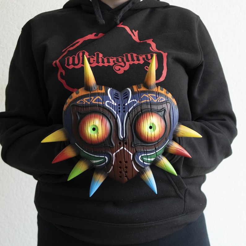 May include: A colorful, detailed, wooden mask with sharp, pointed spikes. The mask is shaped like a heart and has large, yellow eyes with green pupils. The mask is held in front of a person wearing a black hoodie with the word "Witchery" printed on it.