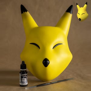 Puede incluir: Una máscara de zorro amarilla con detalles negros en las orejas y la nariz. Una pequeña cabeza de zorro en 3D es visible. Una botella de pintura negra para modelos y un pincel fino también están presentes.