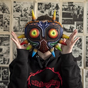 May include: A person wearing a black hoodie with the text "Witchgallery" printed on the front. The person is wearing a large, colorful, heart-shaped mask with sharp points and glowing eyes.