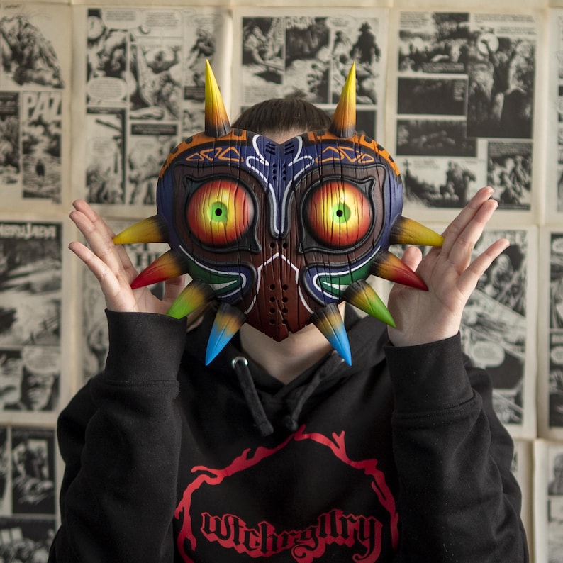 May include: A person wearing a black hoodie with the text "Witchgallery" printed on the front. The person is wearing a large, colorful, heart-shaped mask with sharp points and glowing eyes.
