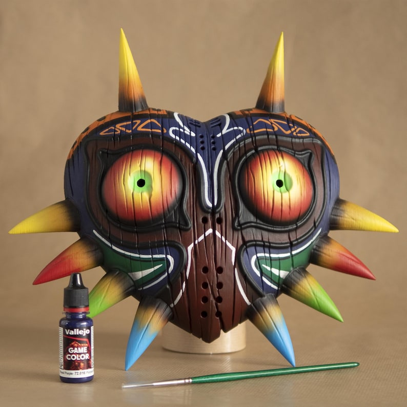 May include: A hand-painted Majora's Mask from The Legend of Zelda video game. The mask is made of clay and has a heart shape with a detailed design. It is painted in shades of brown, red, yellow, green, and blue. The mask is sitting on a wooden base and there is a small bottle of paint and a paintbrush in the foreground.