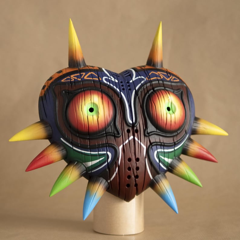 May include: A hand-painted wooden mask with a heart shape and a variety of colors, including red, yellow, green, blue, and orange. The mask has a sharp, pointed design with spikes around the edges. The mask has two large, round eyes with green pupils.