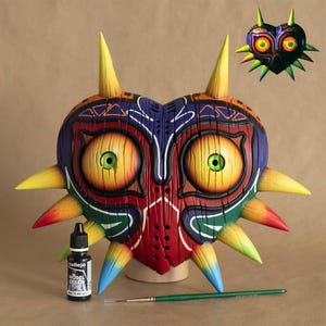 Könnte beinhalten: Eine große, herzförmige Maske mit einem detaillierten, farbenfrohen Design. Die Maske weist markante gelbe Stacheln, große gelbe Augen mit grünen Iris und ein strukturiertes Holz-Aussehen auf. Eine kleinere Version der Maske ist ebenfalls zu sehen, zusammen mit einer Flasche Vallejo Model Color Farbe und einem Pinsel.