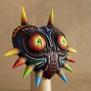 May include: A hand-painted, wooden mask in the shape of a heart with a tribal design. The mask has large, red eyes with green pupils and a series of colorful spikes around the edges.