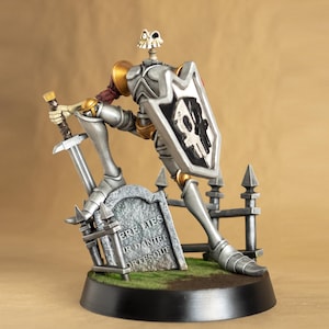 Puede incluir: Una figura de caballero esquelético de plata y oro que sostiene una espada y un escudo. El caballero está de pie sobre una pequeña base con una lápida que dice "Aquí Yace Sir Daniel Ortesque".