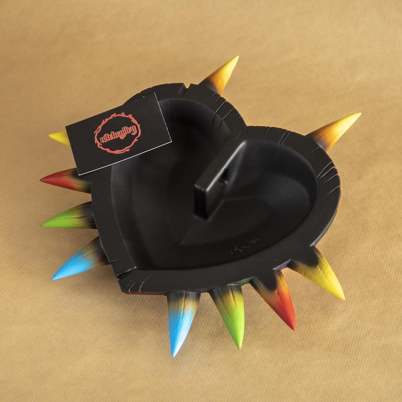 May include: Black heart-shaped dish with a handle and colorful spikes. The dish is made of resin and has a textured surface. The spikes are painted in shades of red, orange, yellow, green, blue, and purple.