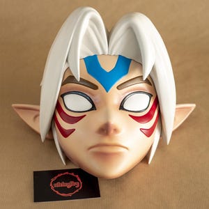 Könnte beinhalten: Eine weiße Maske mit roten und blauen Maldetails. Die Maske zeigt einen Charakter mit weißen Augen, einem blauen Stirnband und roten Markierungen auf den Wangen. Die Maske ist aus einem glatten, harten Material gefertigt.