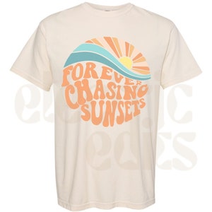 Pu&ograve; includere: Una maglietta beige con una grafica di un tramonto e onde in arancione, giallo, blu e bianco. Il testo "FOREVER CHASING SUNSETS" &egrave; in arancione.
