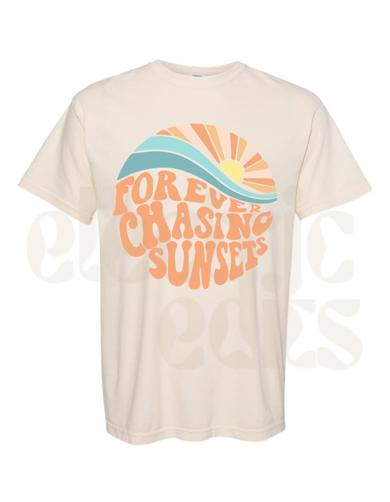 Pu&ograve; includere: Una maglietta beige con una grafica di un tramonto e onde in arancione, giallo, blu e bianco. Il testo "FOREVER CHASING SUNSETS" &egrave; in arancione.