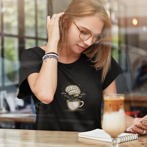 Op de afbeelding: Een zwart T-shirt met een afbeelding van een brein in een koffiekopje. Het brein wordt omringd door sterren en wolken. De persoon die het shirt draagt, zit aan een tafel met een notitieboekje en een glas ijskoffie.