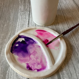May include: A white ceramic watercolor palette with three wells. The wells are filled with pink and purple watercolor paint. A paintbrush is resting on the edge of the palette.