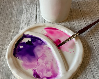 May include: A white ceramic watercolor palette with three wells. The wells are filled with pink and purple watercolor paint. A paintbrush is resting on the edge of the palette.