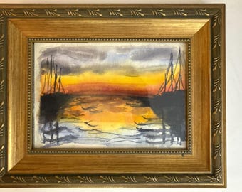 May include: Watercolor painting of a sunset over water, framed in a gold-colored ornate frame. The artwork features a vibrant orange and yellow sky with dark silhouettes of structures and birds. The frame has a detailed, decorative design.