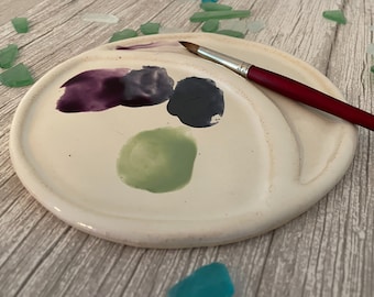 May include: A white ceramic paint palette with three paint wells. The wells contain purple, gray, and green paint. A red handled paintbrush is lying on the palette.