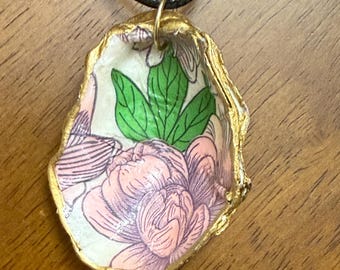 May include: A gold-painted oyster shell pendant necklace with a floral pattern inside. The pattern features pink and green flowers on a white background.