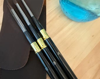 May include: A set of three artist paint brushes with black handles, gold ferrules, and numbered sizes 4, 8, and 12. The brushes are displayed with a dark brown leather case and a blue glass jar.
