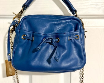May include: A blue leather handbag with a drawstring closure and a top handle. The bag has a gold-tone chain strap and hardware. A small tag hangs from the chain. The bag is hanging from a silver hook on a white door.