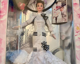 May include: A Barbie doll in a white lace gown with black accents, a wide-brimmed hat, and a parasol. The doll is packaged in a box with a movie poster of "My Fair Lady". The hat has a black and white striped design.