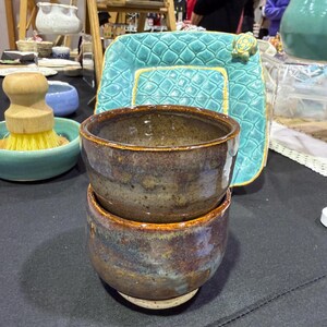 May include: Two handmade ceramic bowls stacked on a white base. The bowls have a mottled brown, blue, and purple glaze with a golden rim. The bowls are part of a collection of handmade pottery.