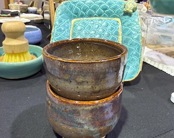May include: Two handmade ceramic bowls stacked on a white base. The bowls have a mottled brown, blue, and purple glaze with a golden rim. The bowls are part of a collection of handmade pottery.