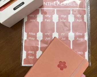 May include: A peach-colored journal with a flower design, a package of monthly labels, and a box of patterned tape. The tape box reads "BASIC PATTERN TAPE" and "rising sun". The journal has a pink elastic band.